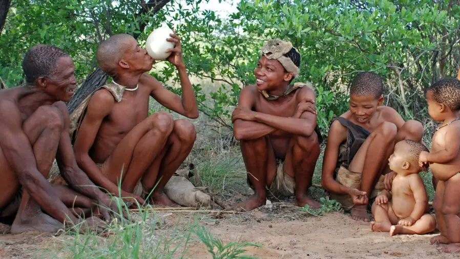 Jóvenes, ancianos, niños, mujeres y hombres de color vestidos con taparrabos, sentados en la tierra.