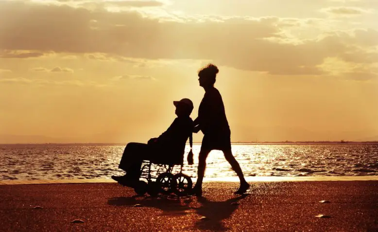 Mujer pasea por la playa a hombre con hemiparesia en silla de ruedas.