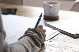 Mesa con taza de cafe, papeles escritos, y mano con lapicera escribiendo un texto narrativo.